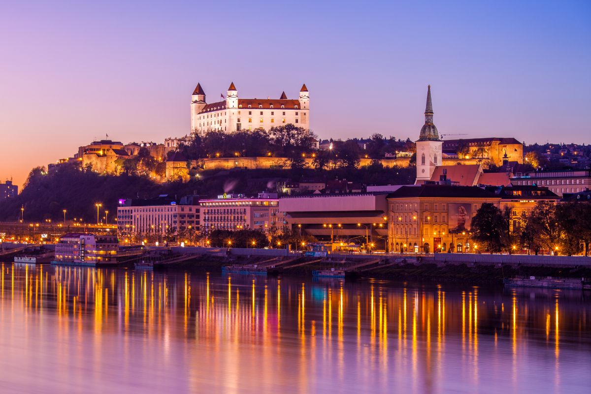 View of Bratislava Castle in the evening from across a river. City lights are reflected in the river.