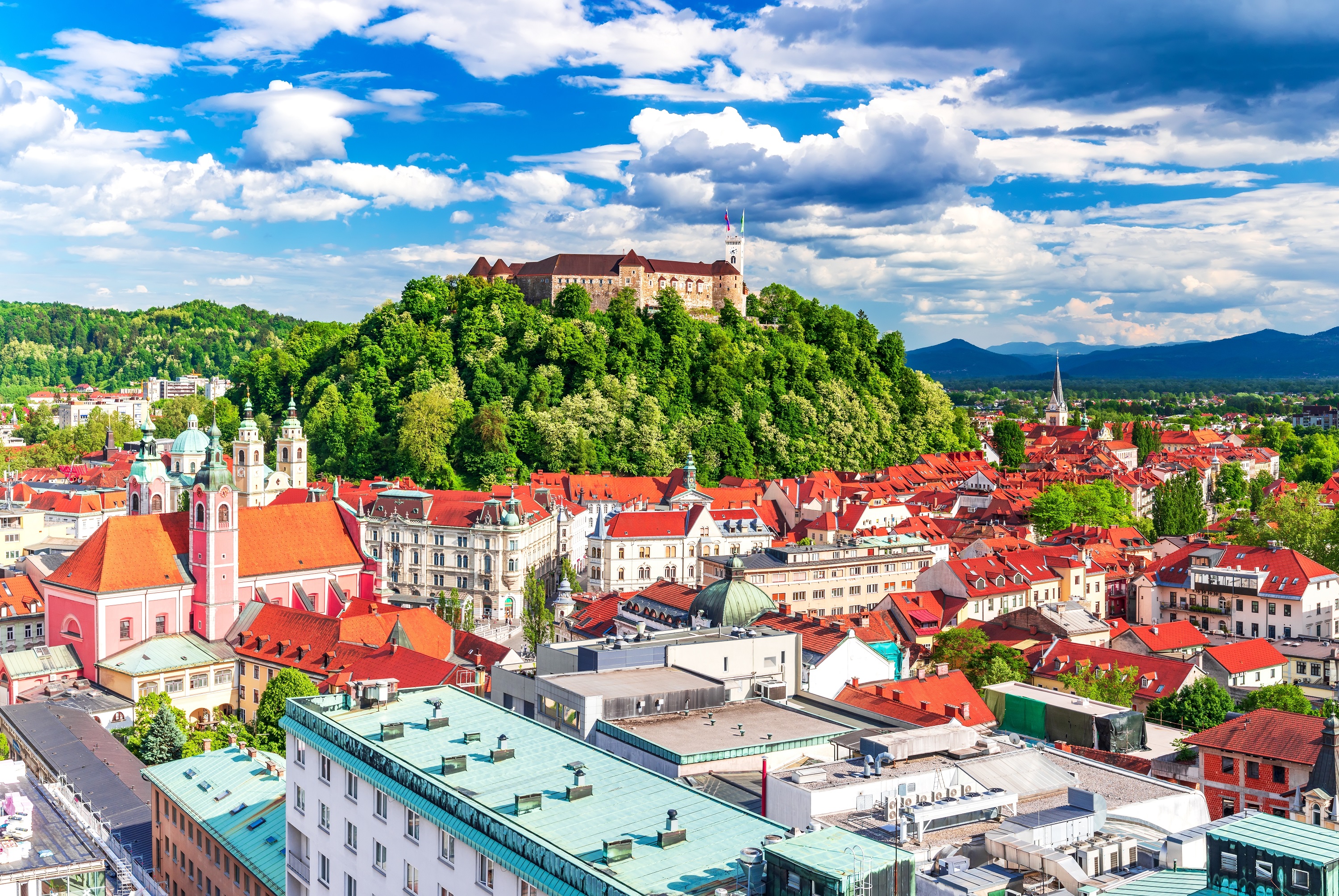 City of Ljubljana, Slovenia. View of city rooftops, mostly terracotta colored. Castle-like building at center of photo on a hill covered in trees. blue sky with white puffy clouds