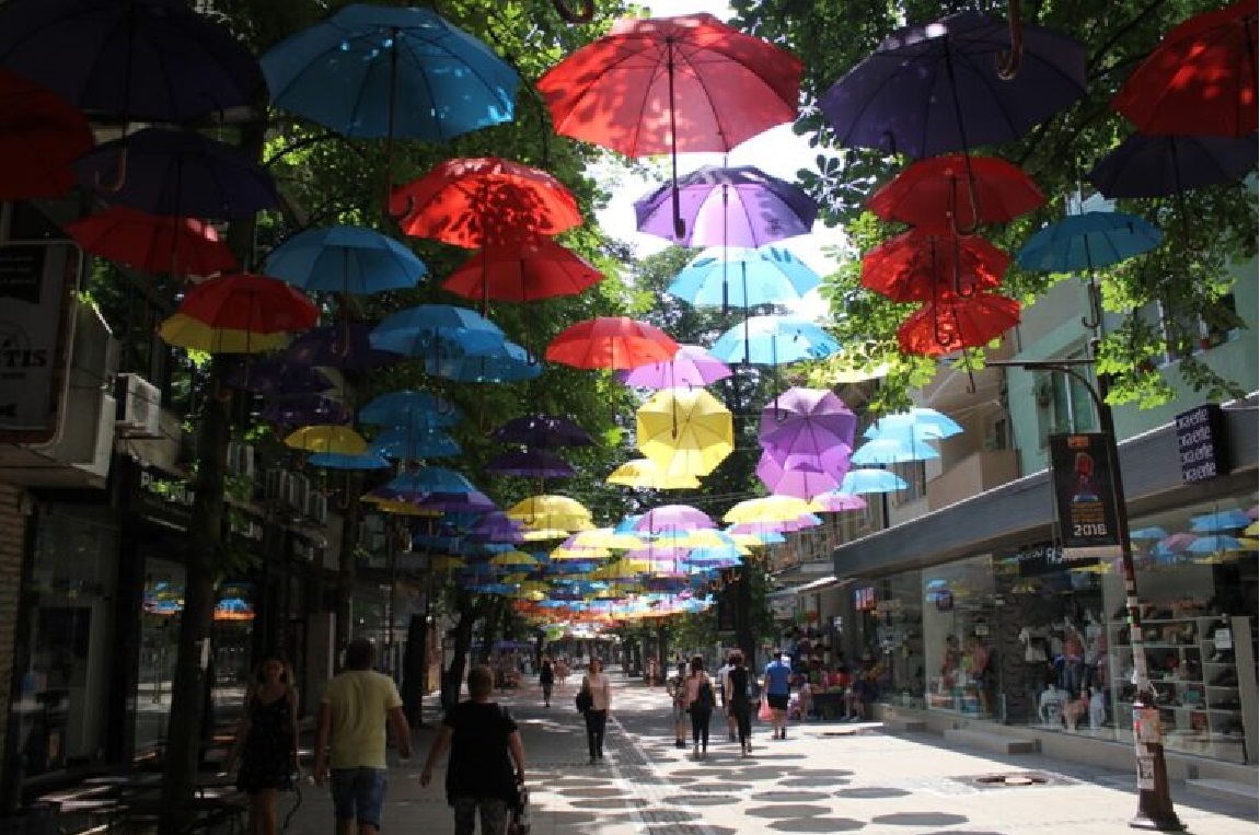 street in Blagoevgrad, Bulgaria. people walking down the street. shops to the right. colorful umbrellas hanging above the street