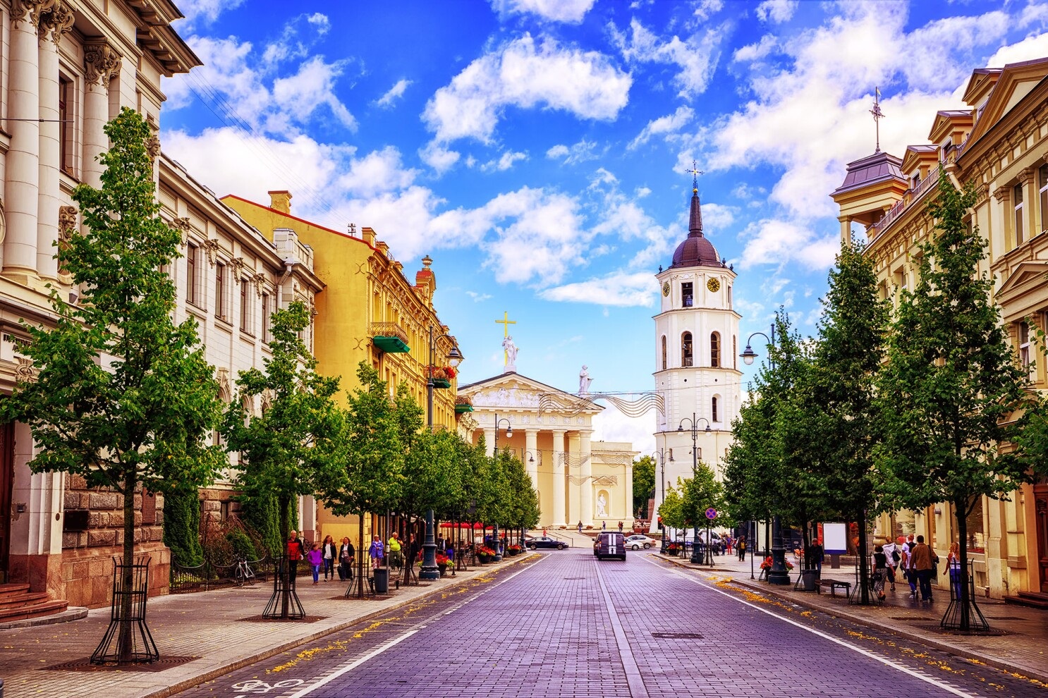 Street view of Vilnius, Lithuania. Buildings on either side of an empty, tree-lined street. There are people on the sidewalks. Blue sky with white fluffy cloud. 