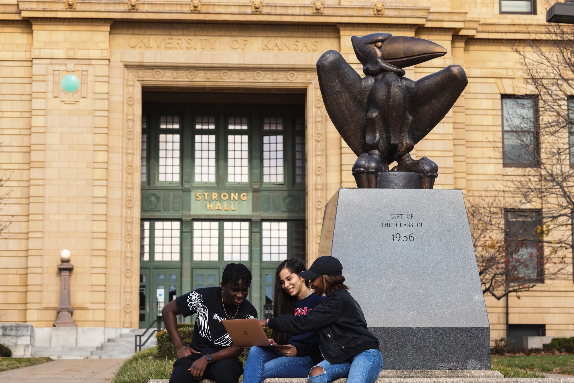 Three students looking at a laptop and smiling while sitting in front of Strong Hall, near a bronze statue of a jayhawk labeled gift of the class of 1956