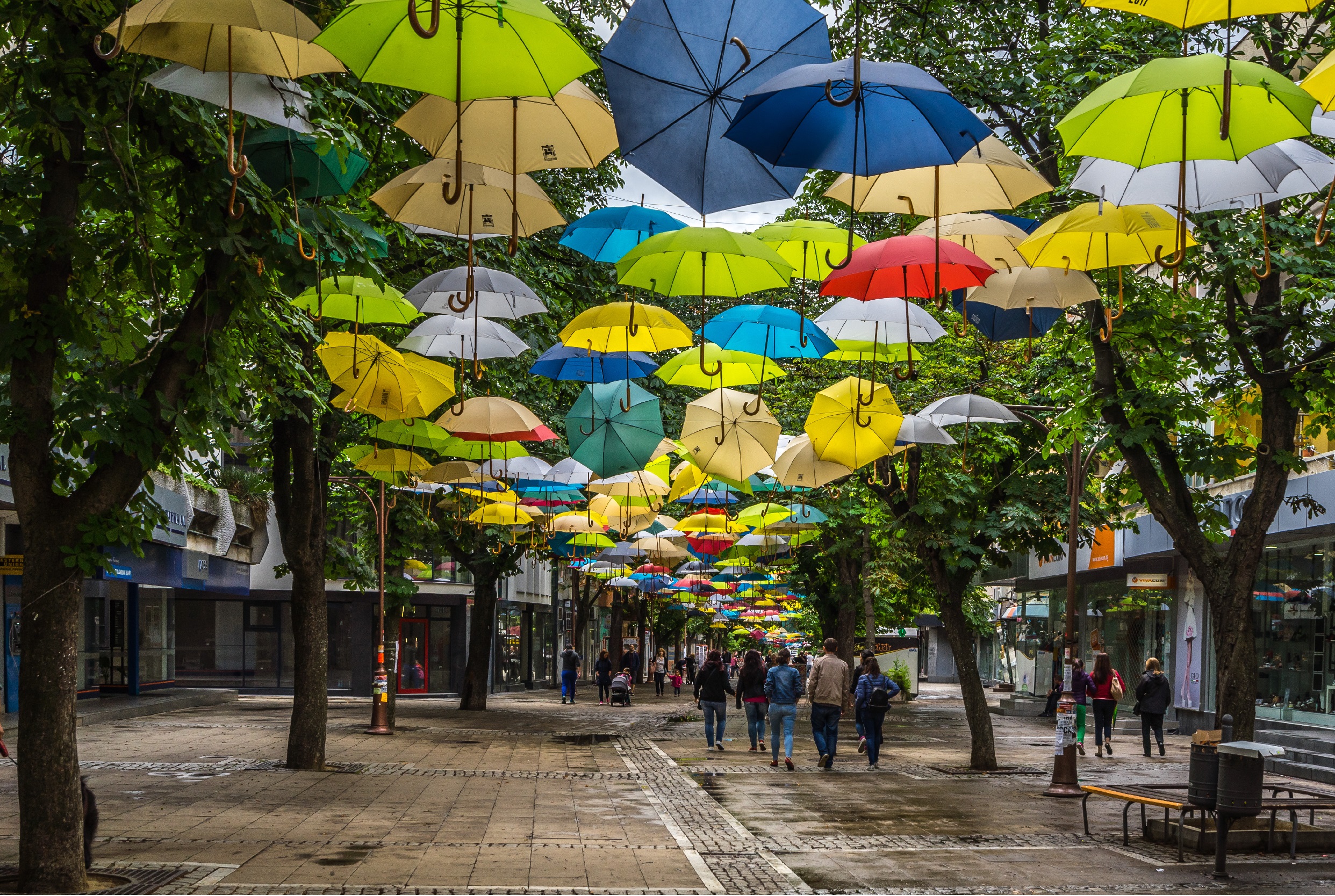 street in Blagoevgrad, Bulgaria. people walking down the street. shops to the right. colorful umbrellas hanging above the street