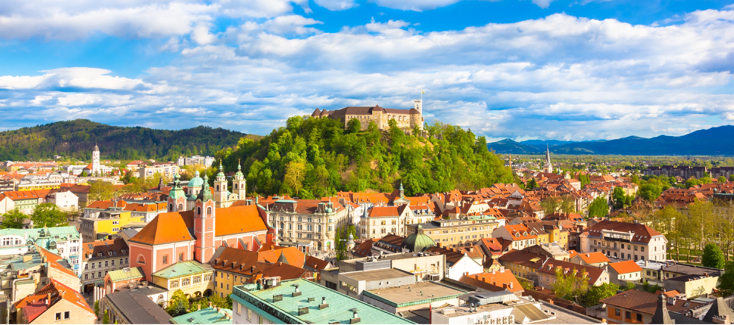 City of Ljubljana, Slovenia. View of city rooftops, mostly terracotta colored. Castle-like building at center of photo on a hill covered in trees. blue sky with white puffy clouds